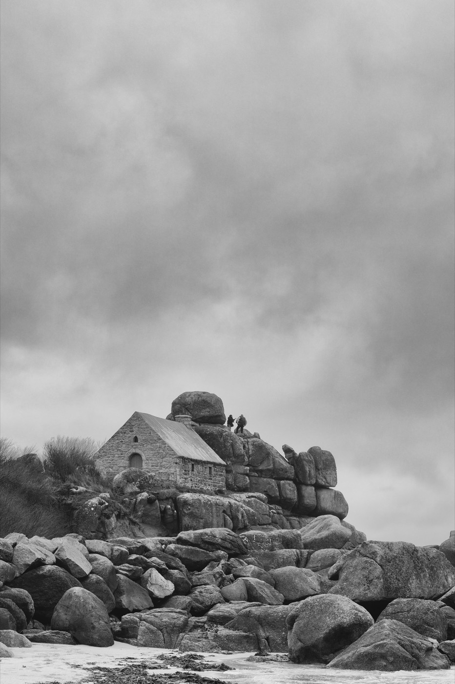 Une petite chapelle de granite tient sur un chaos de blocs, au-dessus de la plage. Deux silhouettes se tiennent près d'elle, à peine plus grandes que les pierres.