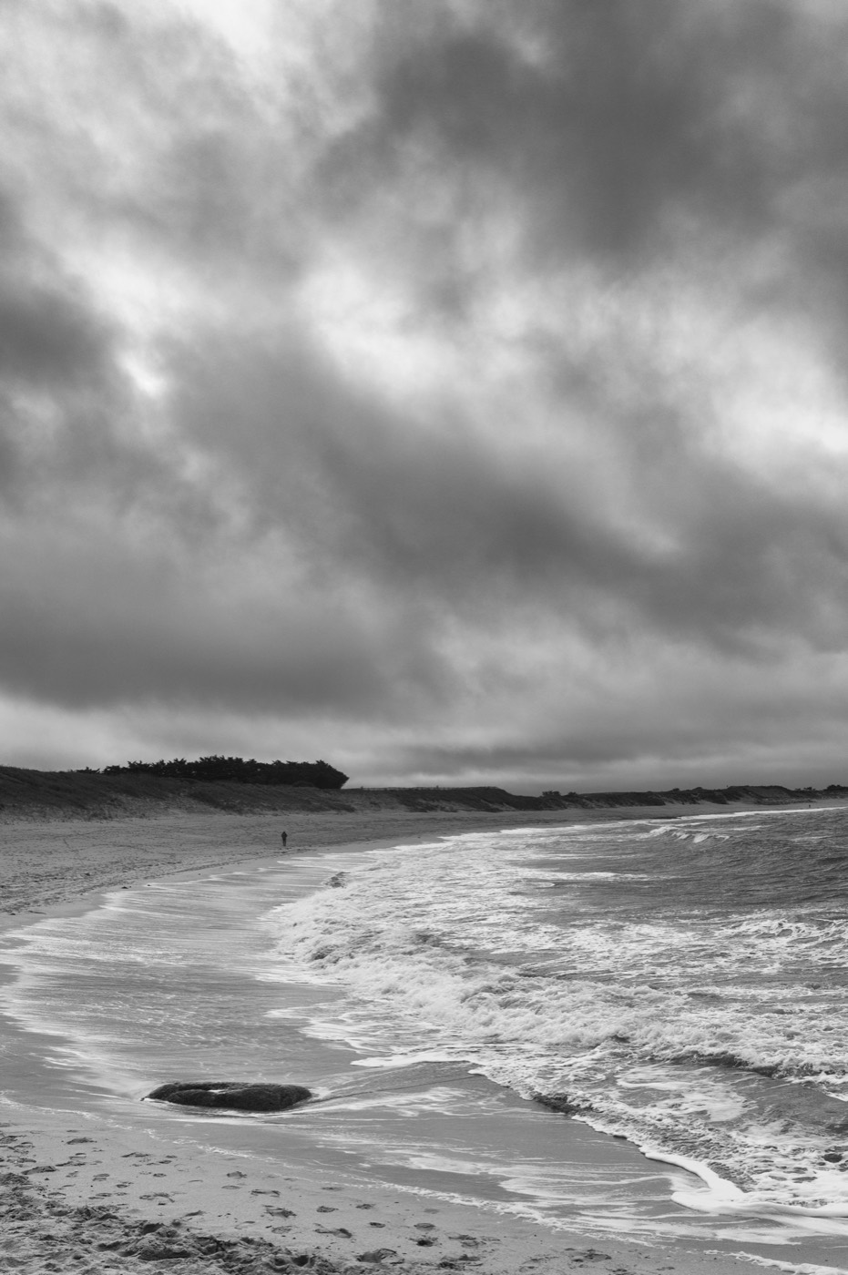 L'écume dessine une longue courbe sur le sable ; une silhouette minuscule marche au bout de la plage. Le ciel est chargé jusqu'à l'horizon.