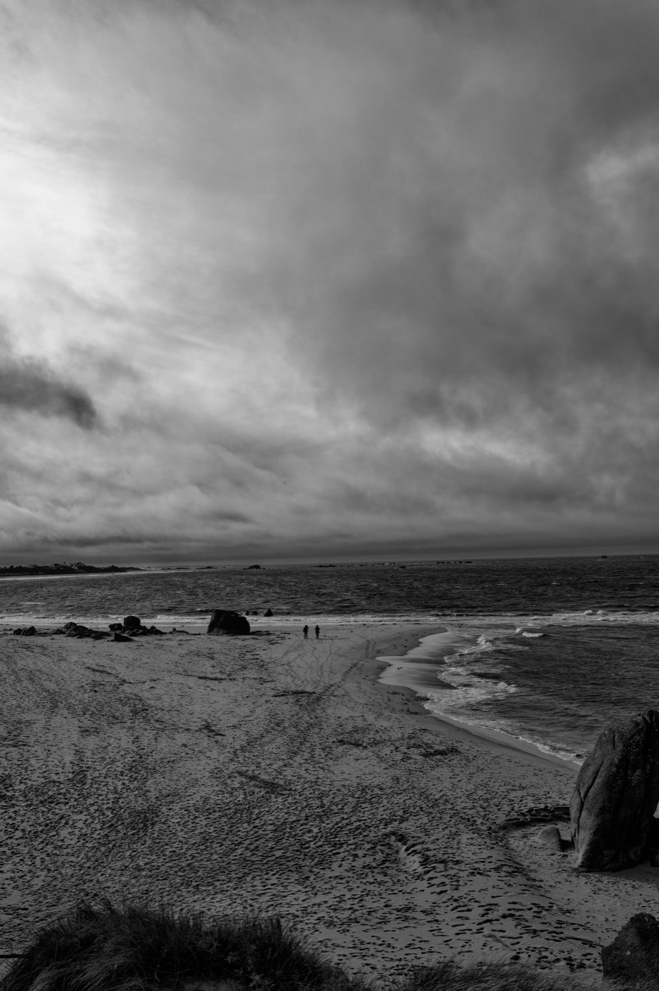 Deux silhouettes minuscules marchent sur l'estran, entre le sable clair et la mer. Un ciel lourd occupe les trois quarts de l'image.