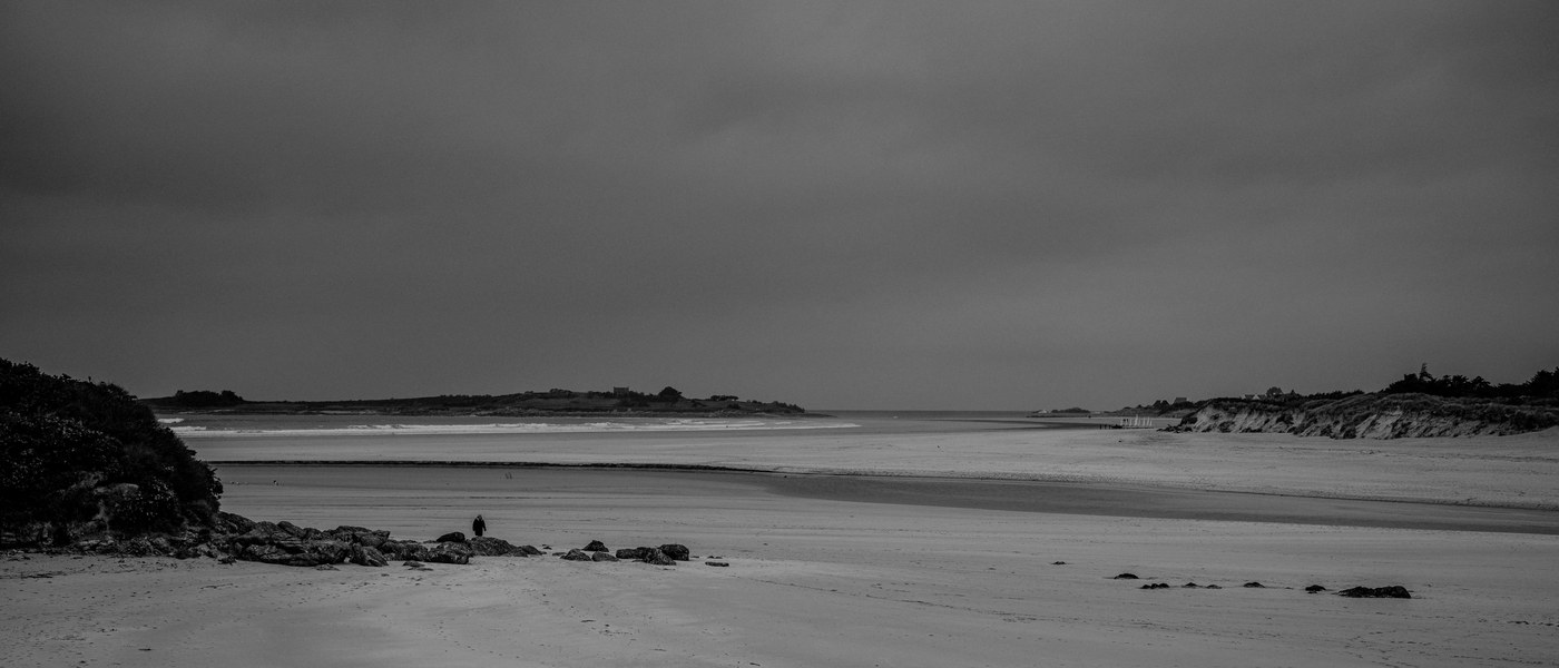 Une grève immense découverte par la marée sous un ciel plombé ; au loin une pointe boisée ferme la baie, et une silhouette seule, minuscule, se tient debout près des rochers.