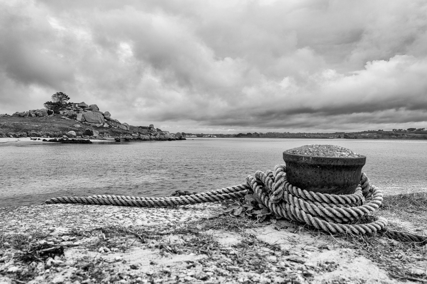 Une bitte d'amarrage de fonte, un cordage lové autour d'elle, posée sur l'estran ; au fond, un îlot boisé sous des nuages.