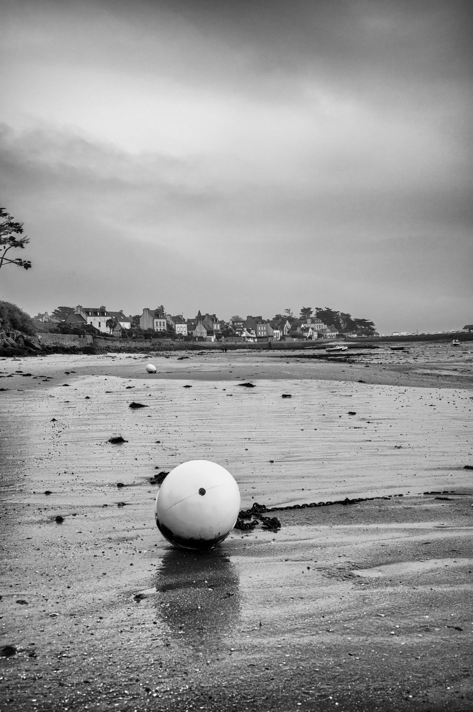 Une bouée blanche et ronde échouée sur le sable humide ; au fond, une côte basse et un ciel voilé.