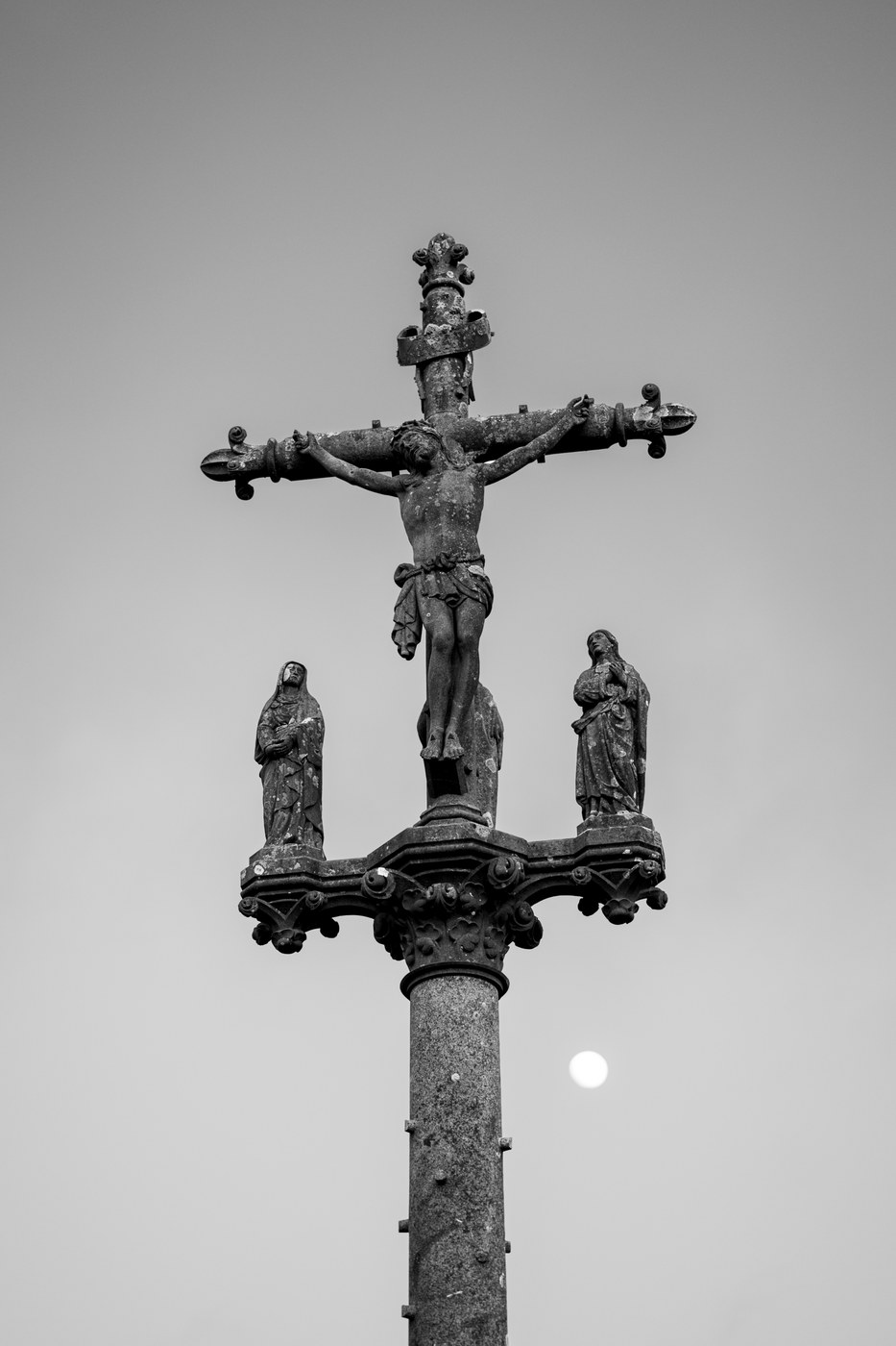 Un calvaire de pierre se détache sur un ciel gris uni ; la lune, minuscule et blanche, flotte à droite du fût.