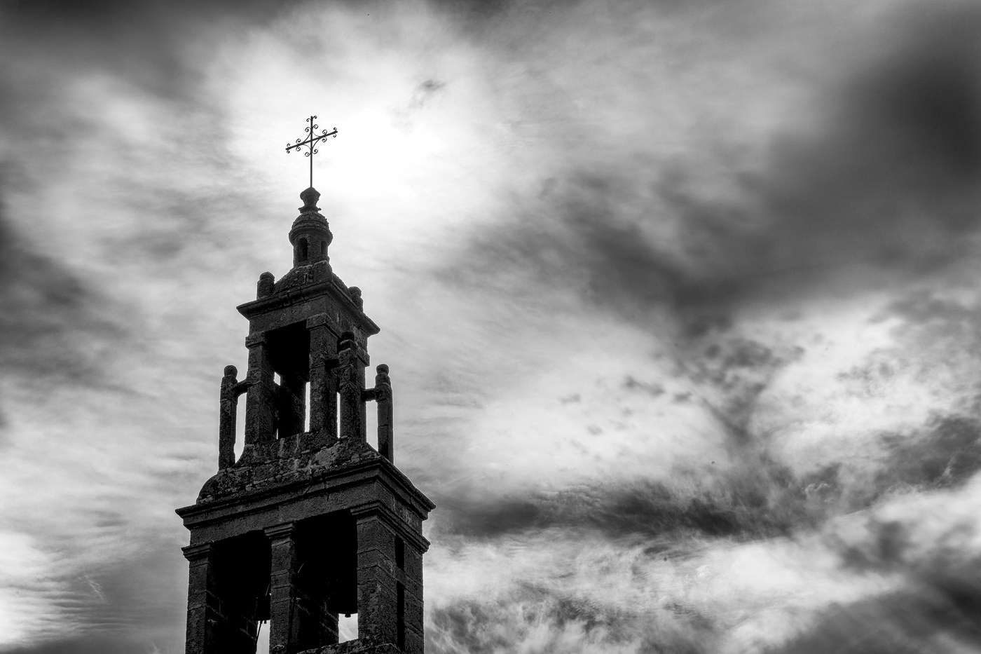 Un clocher de pierre en silhouette ; le soleil est exactement derrière la croix du sommet et les nuages tournent autour.