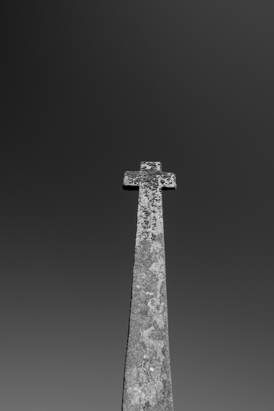 Une croix de granite vue en contre-plongée, la pierre couverte de lichen, sur un ciel sombre qui s'éclaircit vers le bas.