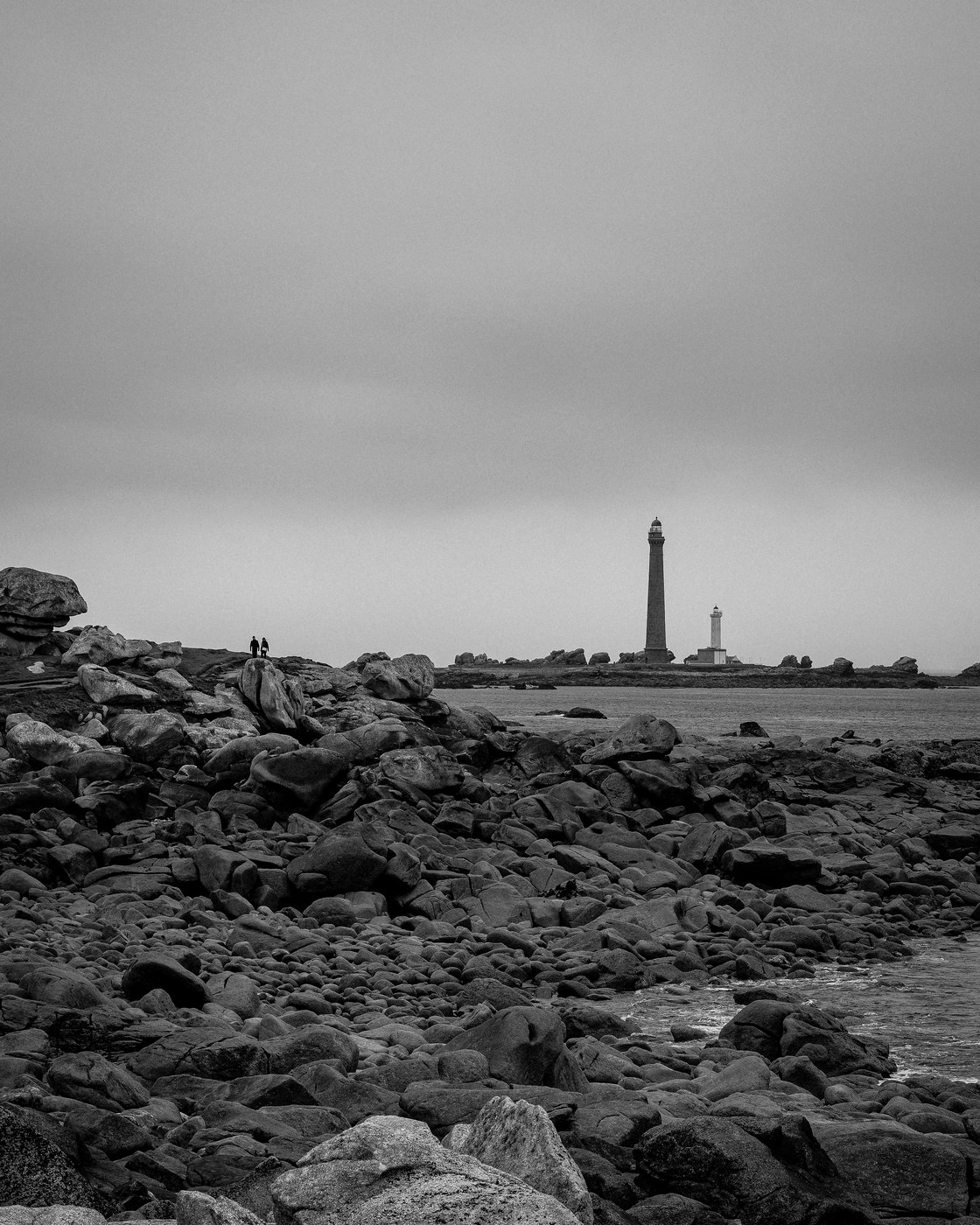 Deux phares se dressent à l'horizon, l'un haut et sombre, l'autre bas et clair ; au premier plan un vaste chaos de blocs de granite, et deux silhouettes minuscules debout sur les rochers, à gauche.