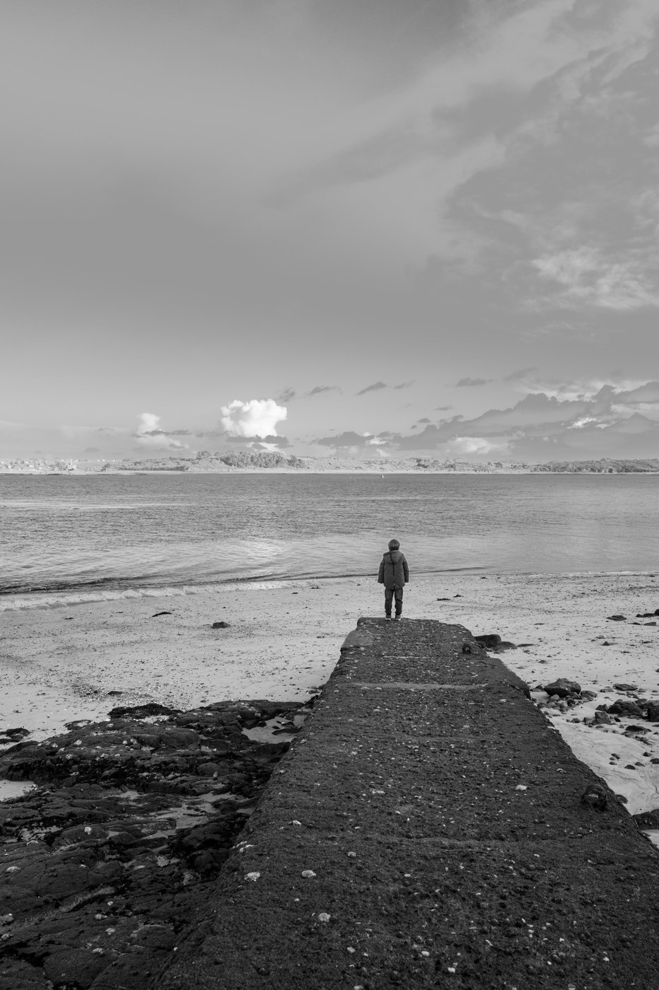 Une silhouette de dos, seule au bout d'une longue jetée de pierre qui s'avance dans l'eau calme ; le ciel est bas et gris.