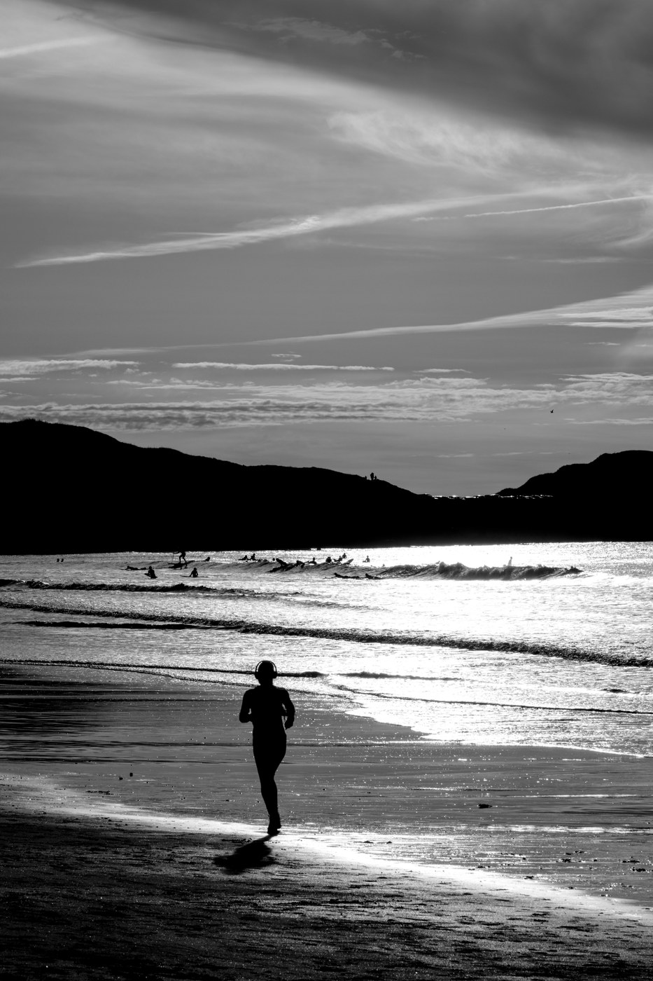 Une silhouette entièrement noire marche sur l'estran à contre-jour ; la lumière se réfléchit en une large bande sur le sable mouillé.