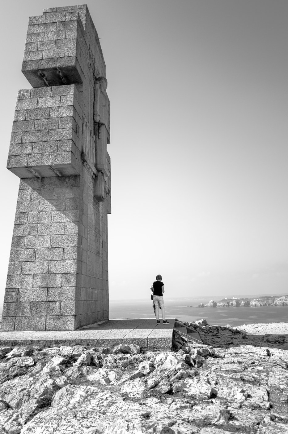 Un monument de granite massif se dresse au bord de la mer ; une silhouette minuscule, de dos, se tient à son pied et regarde le large.