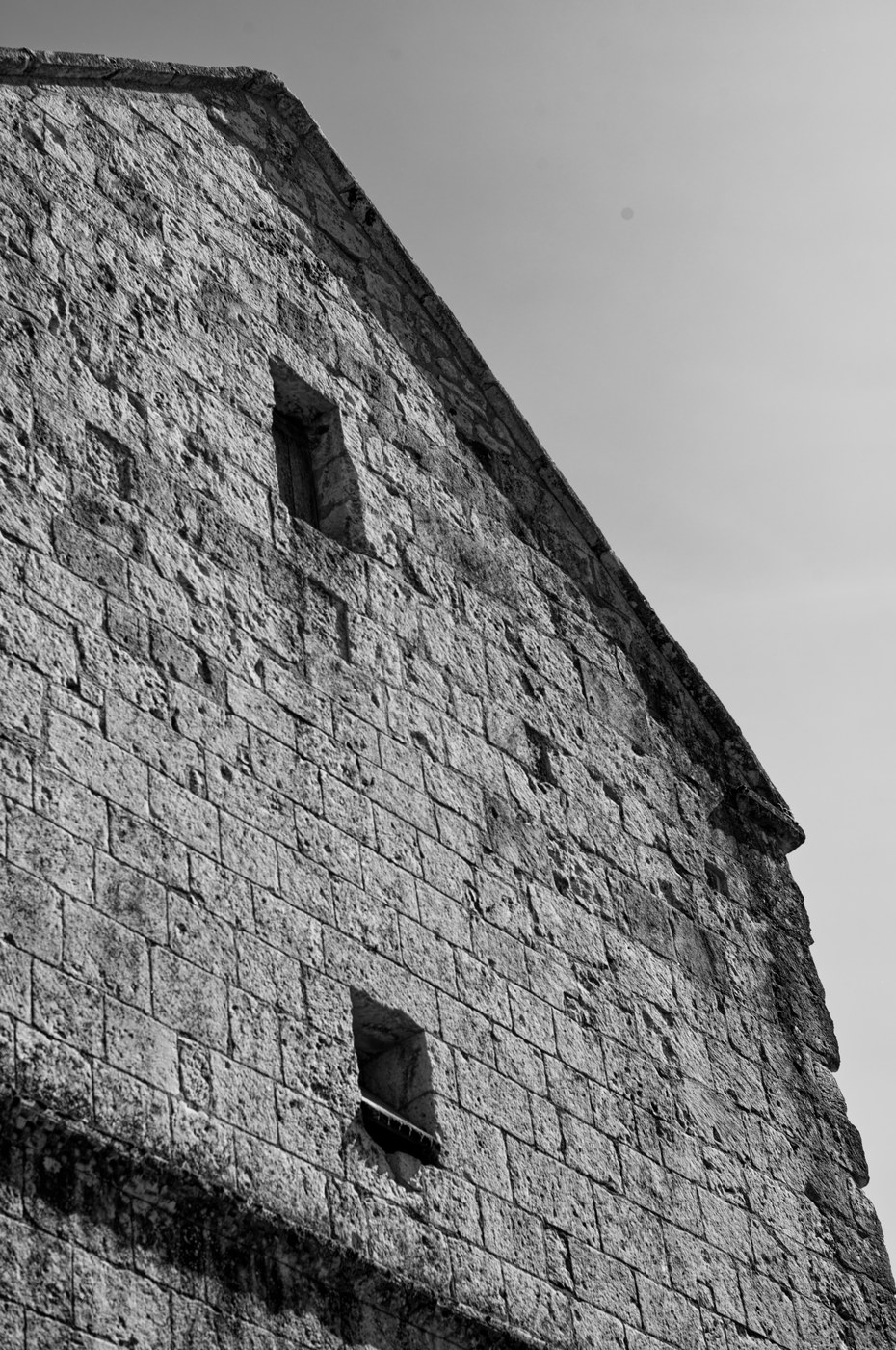 Un mur de pierre vu en contre-plongée, deux ouvertures étroites percées dans l'appareil, le ciel au-dessus.