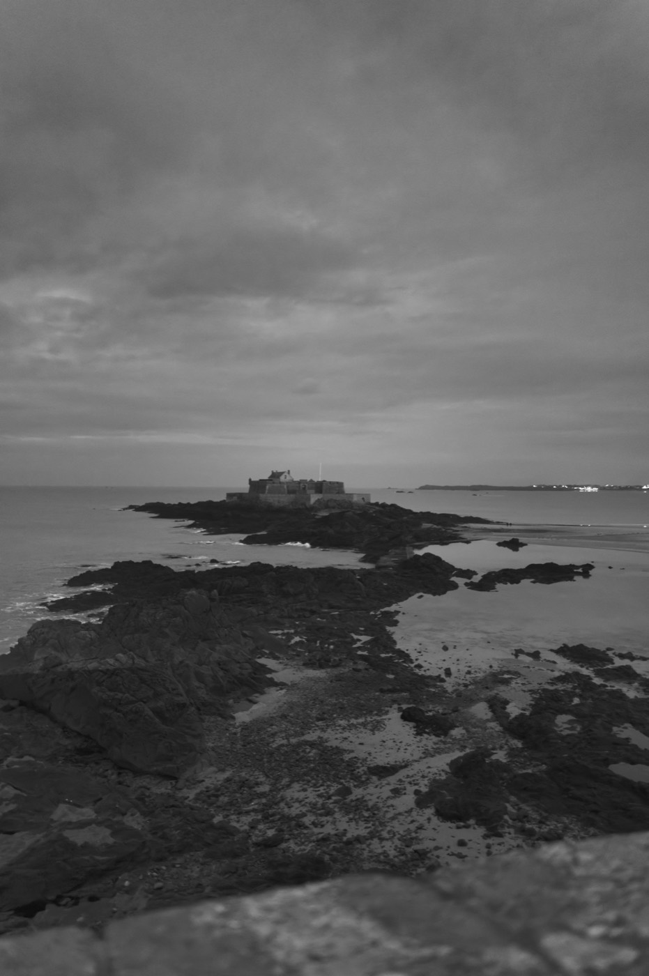 Un fort posé sur un îlot rocheux à la tombée du jour, relié à la côte par l'estran découvert. Les lumières de la ville s'allument au loin.