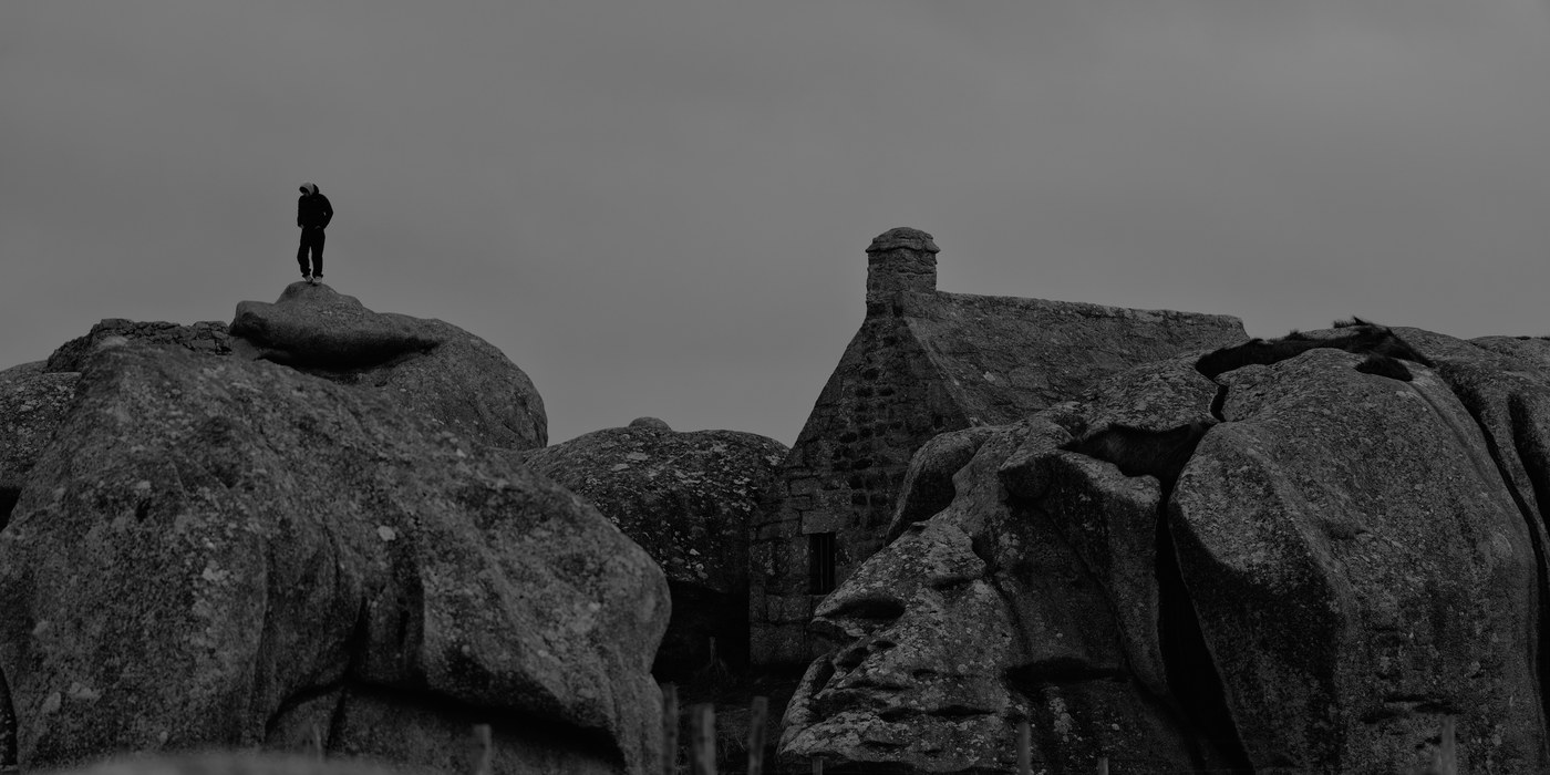 Une silhouette solitaire, capuche relevée, se tient debout au sommet d'un rocher de granite ; en dessous, un chaos de blocs et le pignon d'une maison de pierre encastrée entre eux ; la moitié haute de l'image est un ciel gris et nu.