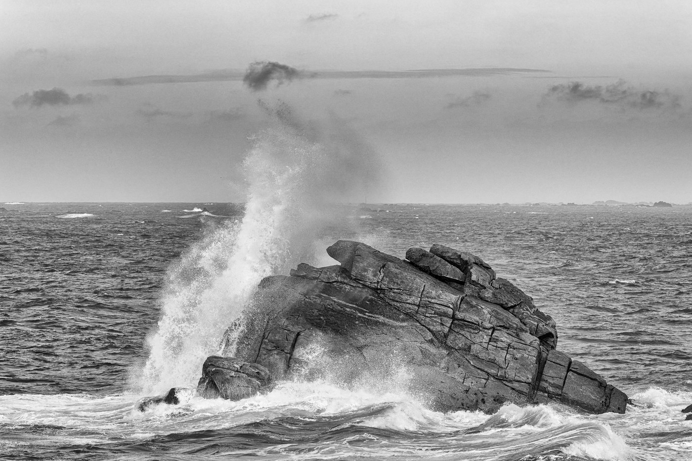 Une vague éclate contre un rocher plat posé dans la mer ; l'écume monte en colonne au-dessus de la pierre.